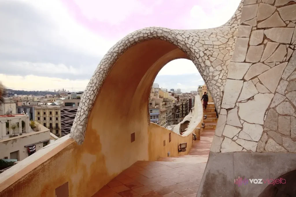 Un arco revestido de trencadís blanco sobre una escalera de la azotea de La Pedrera, con los tejados del Eixample al fondo