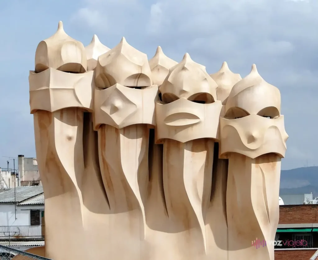 Un grupo de 7 chimeneas de color crema en la azotea de La Pedrera, cada una con forma de cabeza con casco y visera, sobre un cielo azul