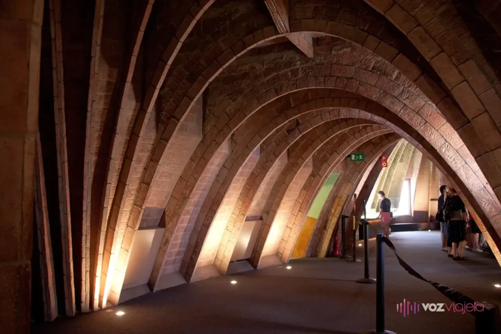 El desván de La Pedrera: un largo túnel de arcos catenarios de ladrillo visto, con luz cálida y visitantes entre las vitrinas de la exposición sobre Gaudí