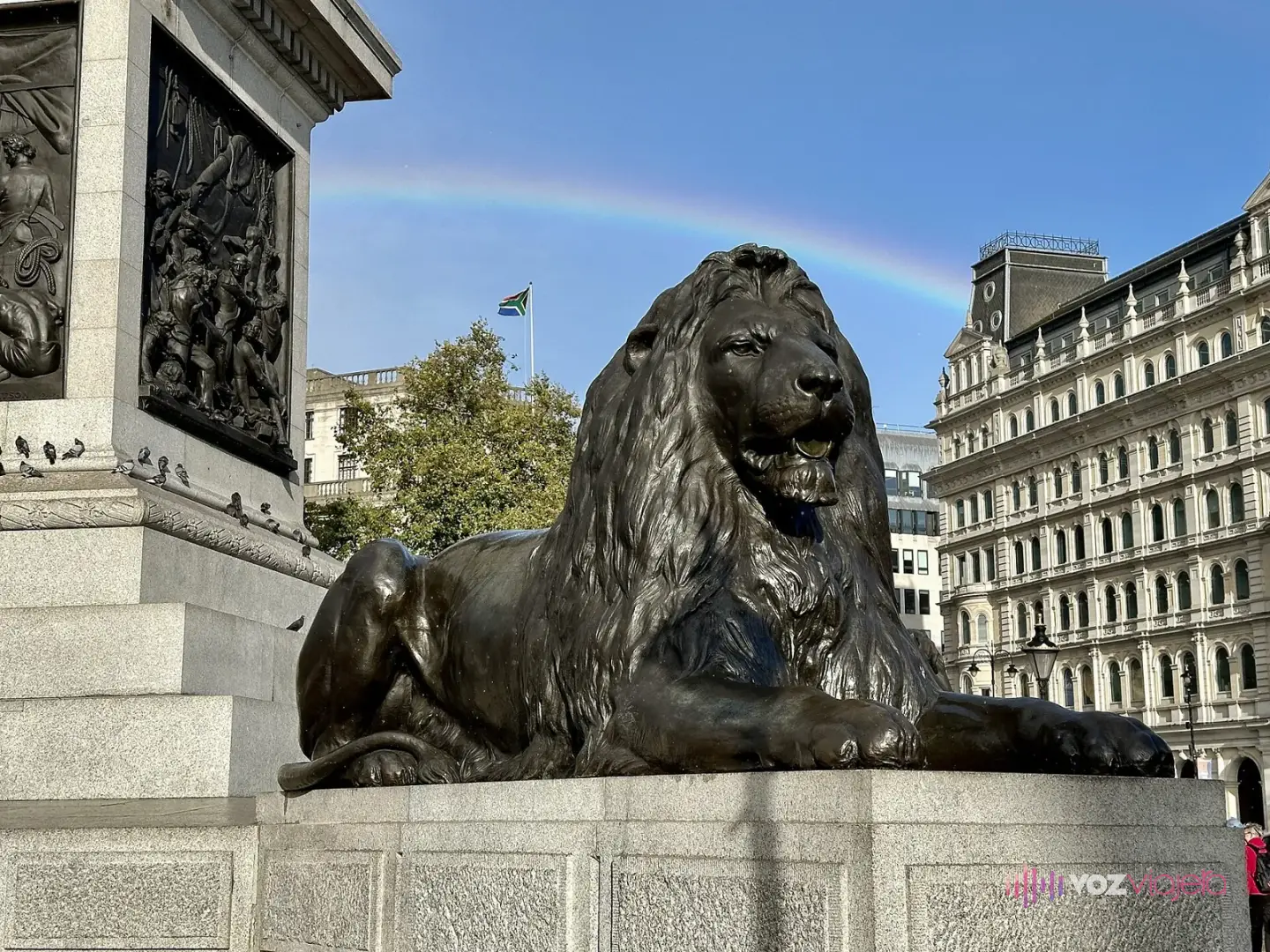 Uno de los 4 leones de bronce de Landseer tumbado al pie de la Columna de Nelson, junto a un relieve de bronce y con un arcoíris en el cielo