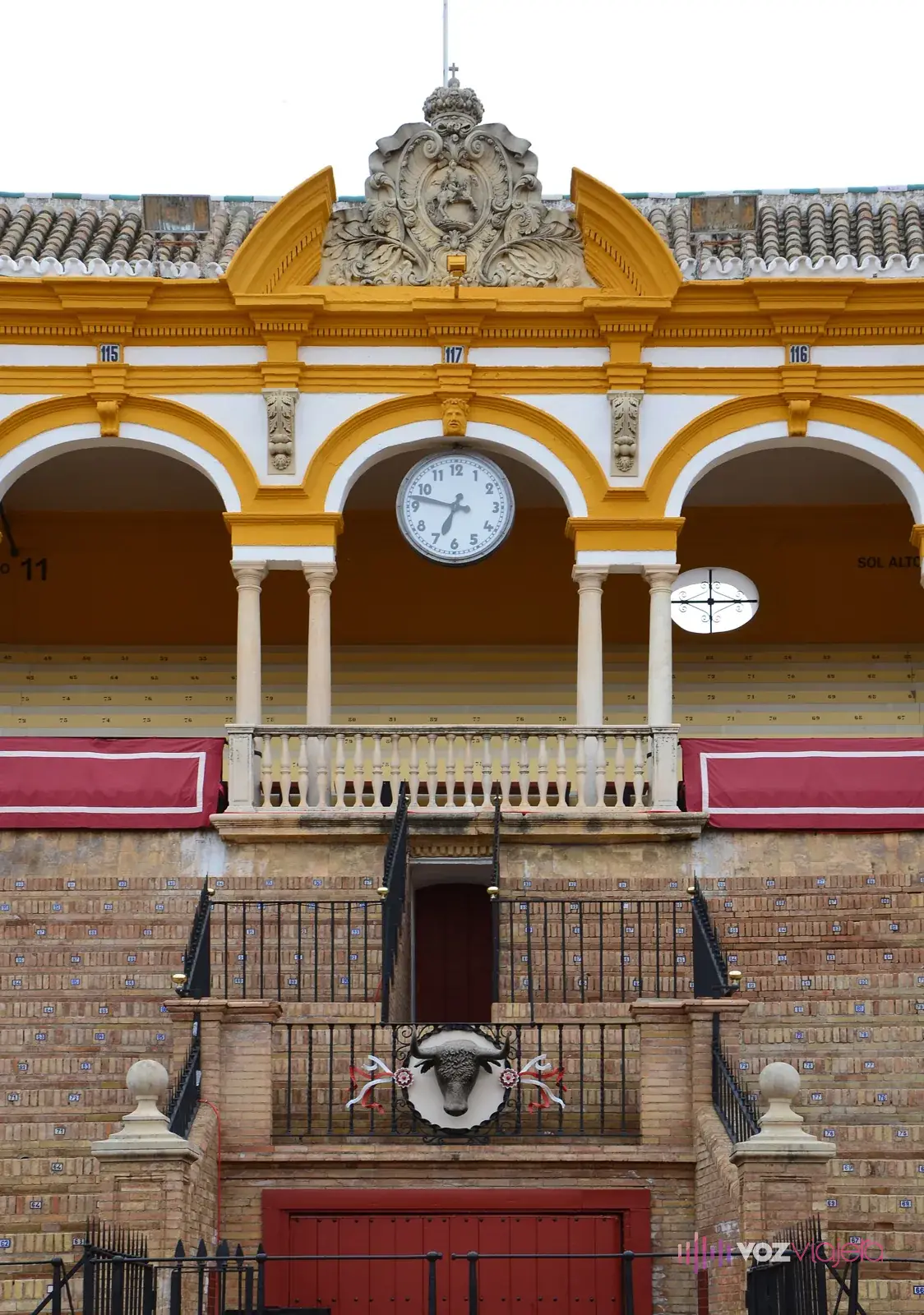 Palco del interior de la plaza de toros de la Maestranza de Sevilla, con arcos amarillos, un reloj y un balcón sobre una puerta roja del ruedo