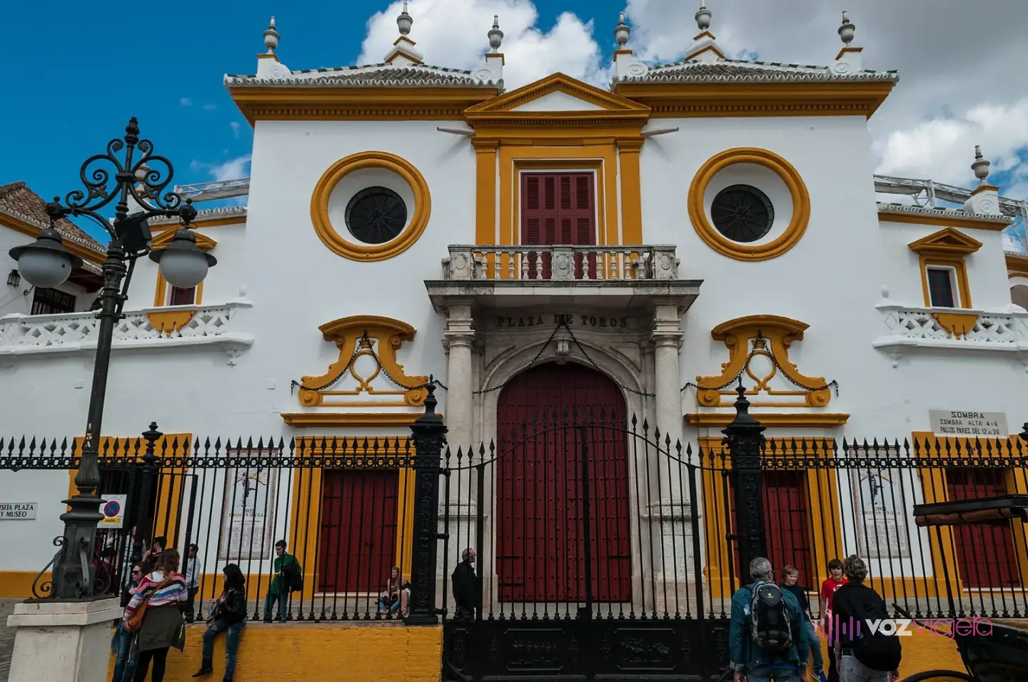 Portada principal de la plaza de toros de la Maestranza de Sevilla: fachada blanca y albero con dos óculos, balcón y puerta roja tras una verja, con visitantes delante