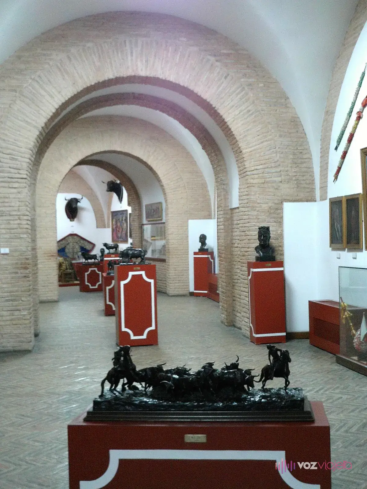 Sala expositiva de la plaza de toros de la Maestranza de Sevilla: galería abovedada de ladrillo con bustos, vitrinas, cuadros y un bronce en primer plano