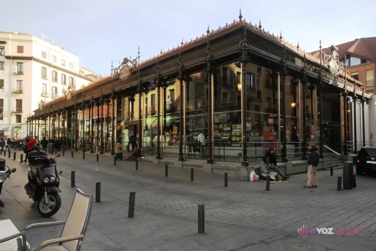 Edificio de hierro y cristal del Mercado de San Miguel en Madrid, con sus columnas de hierro oscuras, la crestería del tejado y la plaza adoquinada delante