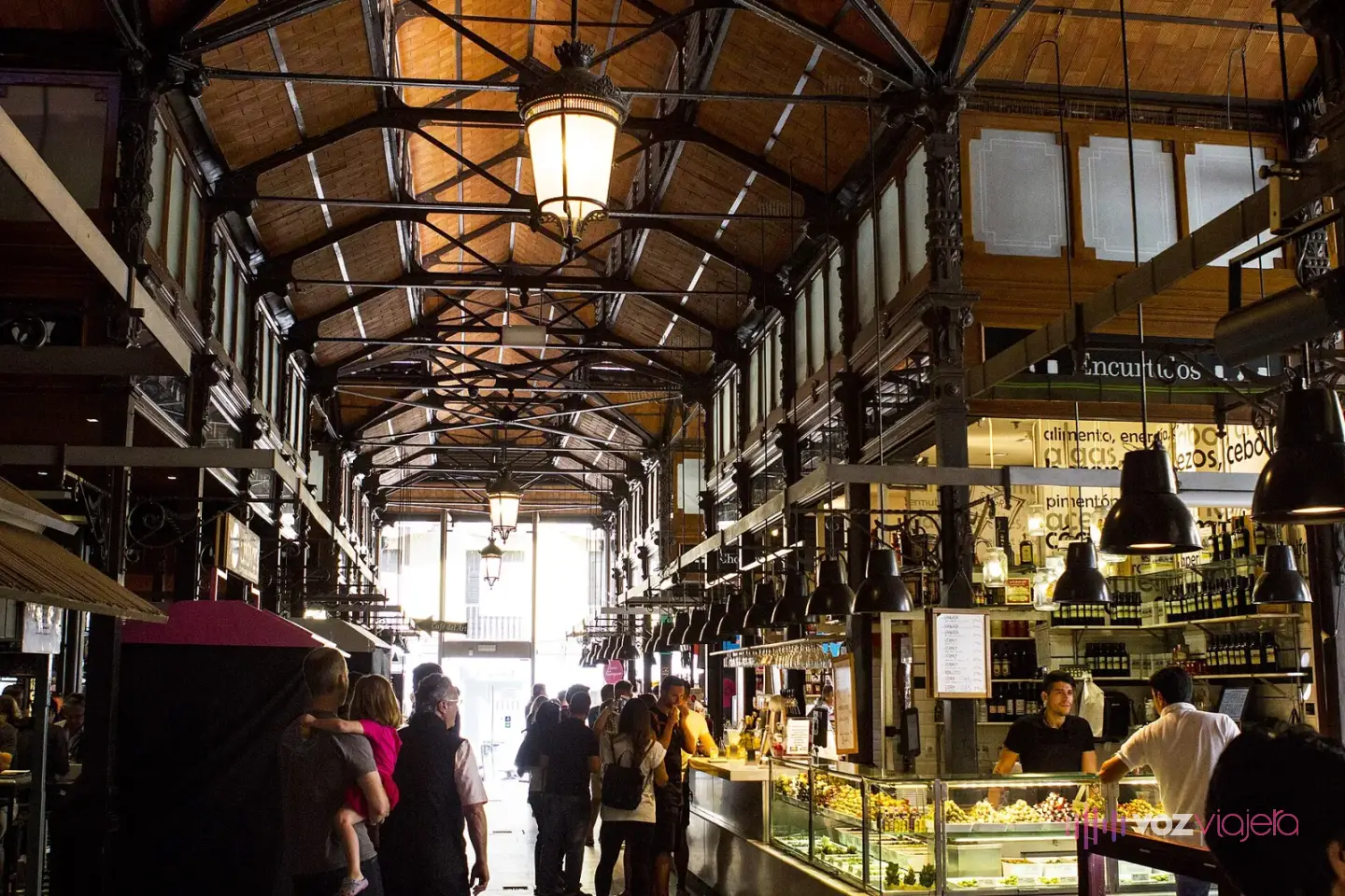 Interior del Mercado de San Miguel: una sola nave con columnas y cerchas de hierro bajo el techo, faroles colgados y puestos con barra a los lados