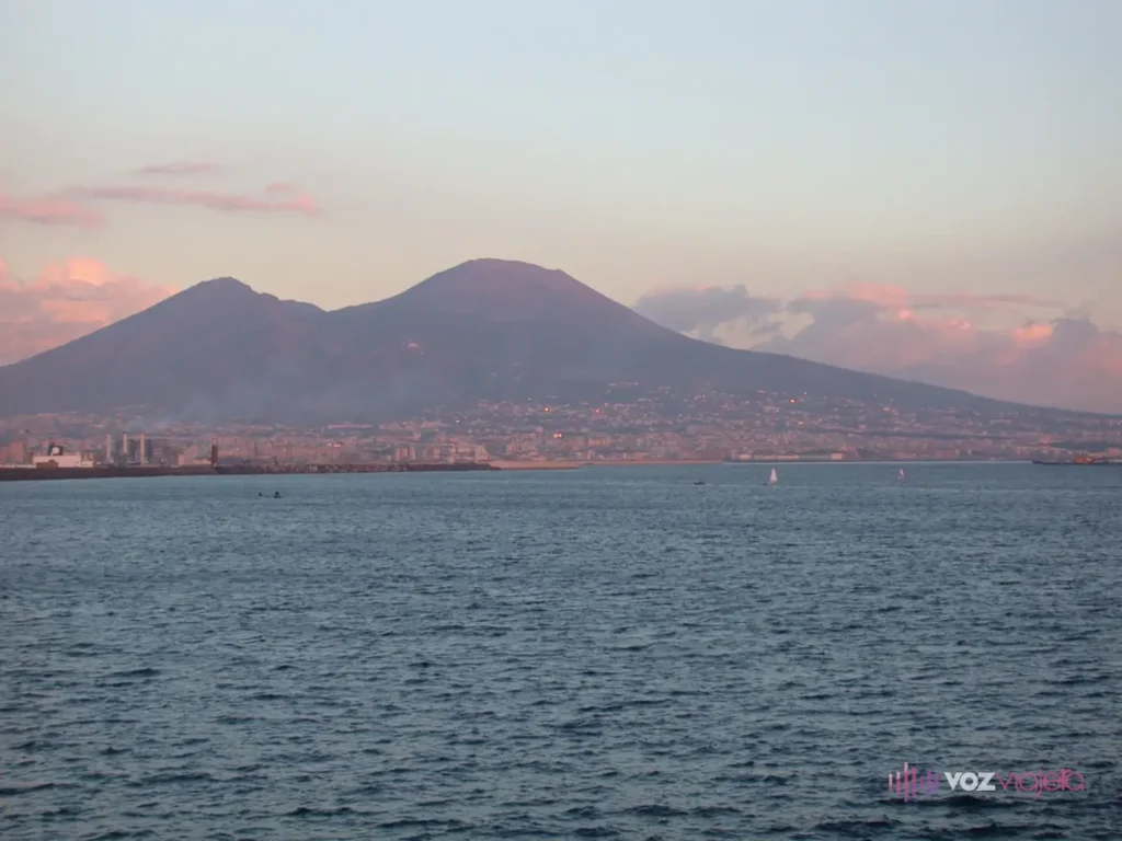 El Vesubio al otro lado de la bahía de Nápoles al atardecer: a la izquierda la cresta baja y dentada del Monte Somma y a la derecha el Gran Cono, más alto y redondeado, sobre una franja de casas y el espigón de un puerto