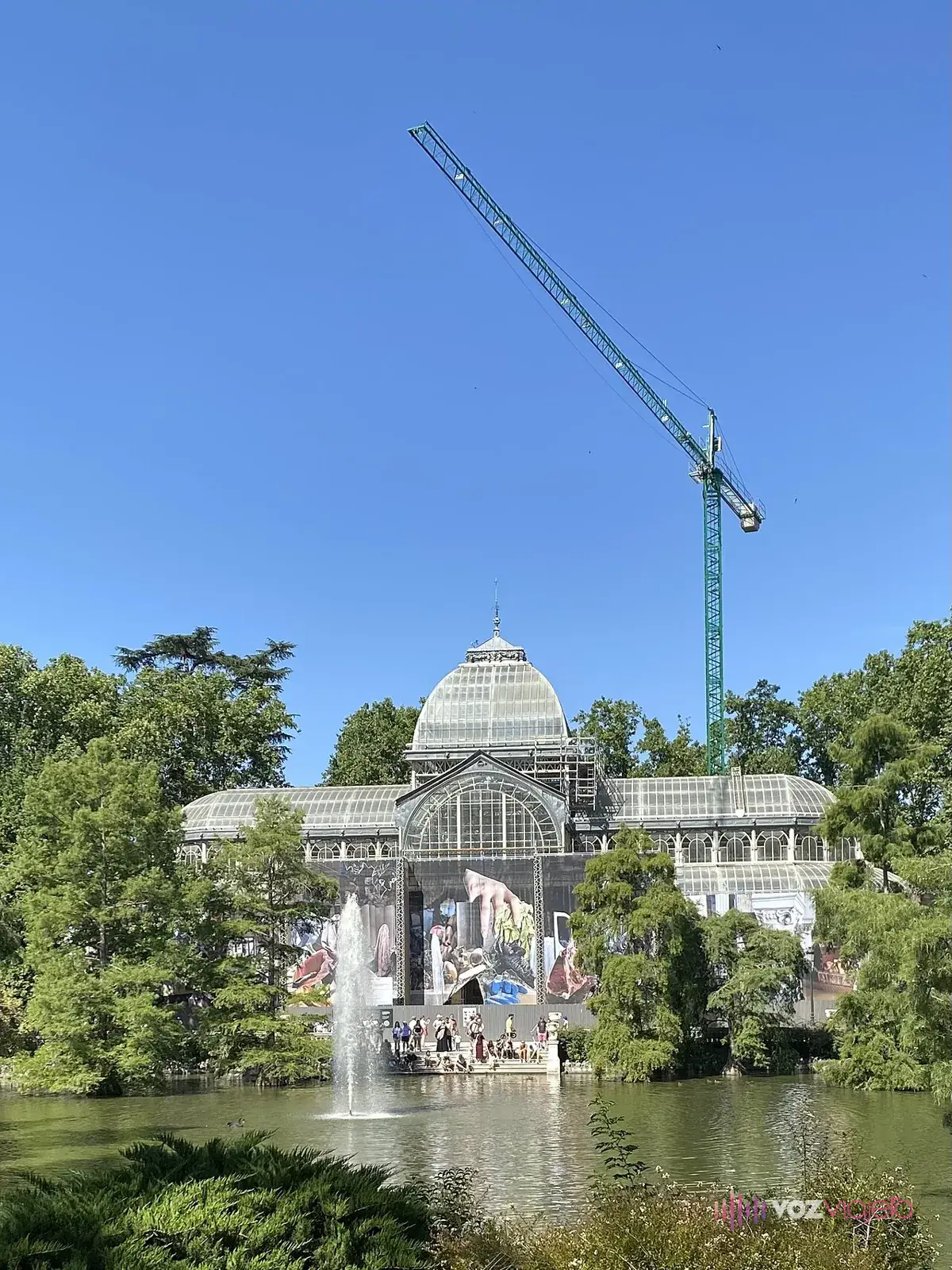 El Palacio de Cristal en obras visto desde el lago: una grúa alta sobre la cúpula y la parte baja cubierta por una lona con imágenes