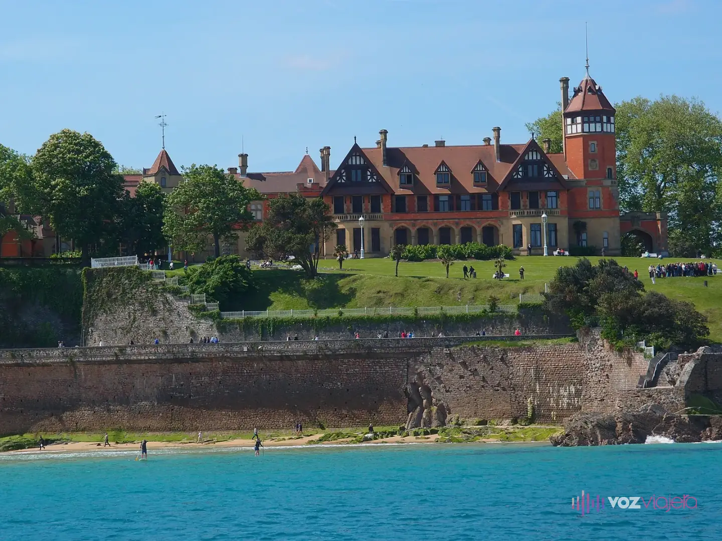 El Palacio de Miramar, de ladrillo rojo con tejados a dos aguas y una torre, sobre los jardines y el muro de piedra del Pico del Loro, junto al mar