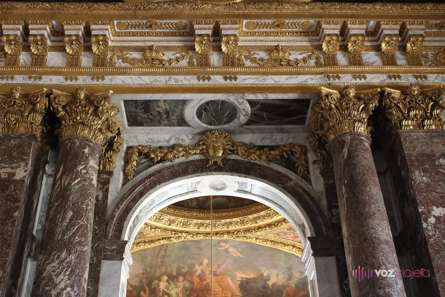 Columnas de mármol rojo con capiteles dorados y un arco bajo un techo pintado, en la Galería de los Espejos de Versalles