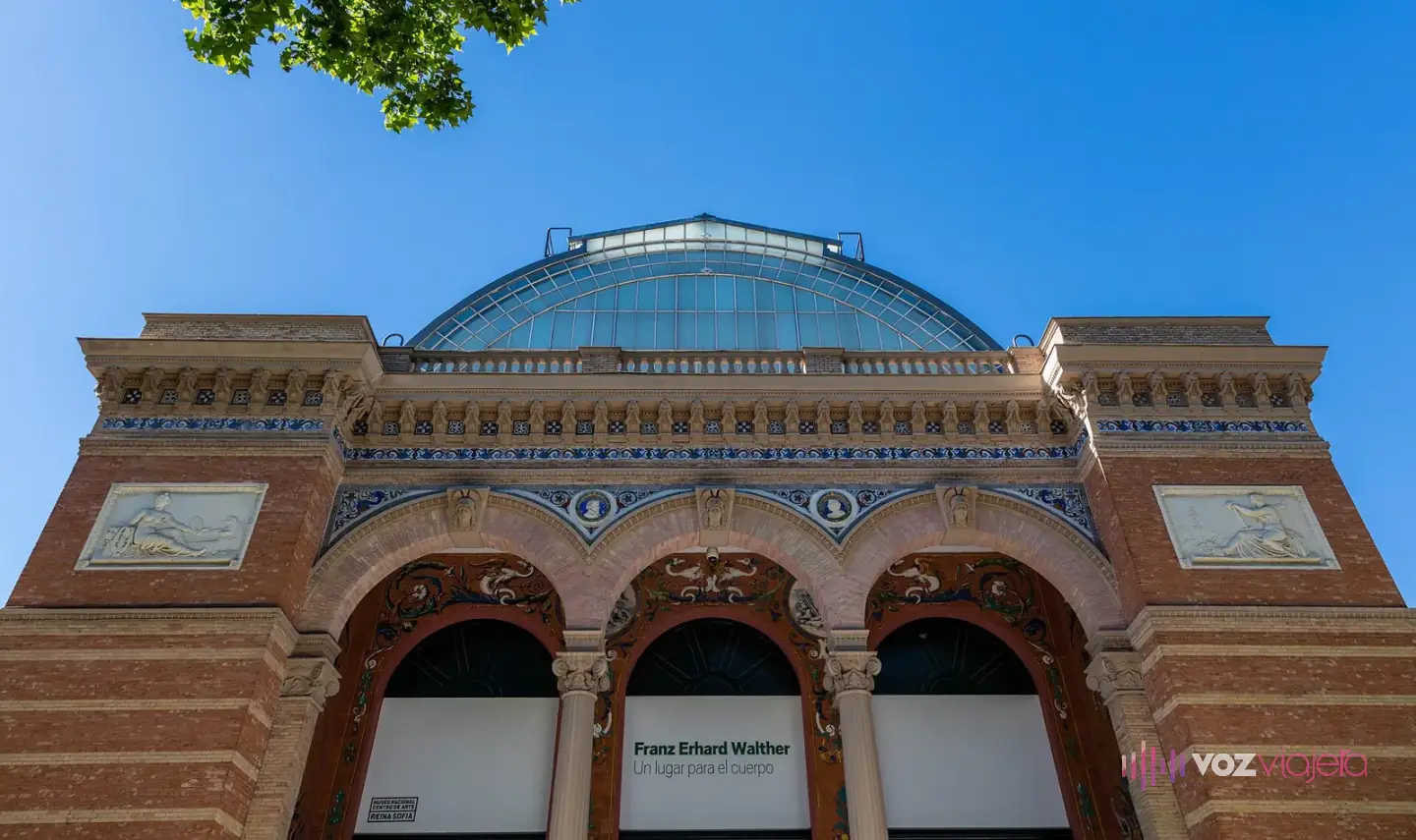 Fachada del Palacio de Velázquez en el Retiro, de ladrillo de dos colores con azulejos, tres arcos de entrada y bóveda de hierro y cristal