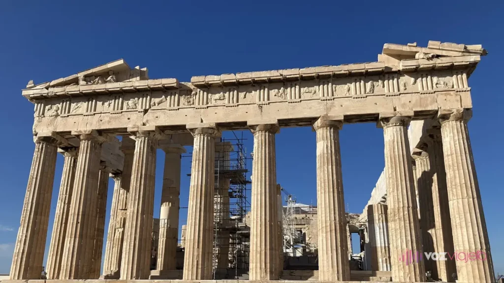 La fachada este del Partenón bajo un cielo azul intenso: 8 columnas dóricas que sostienen el entablamento y los restos del frontón, con una torre de andamio dentro del templo, detrás de las columnas