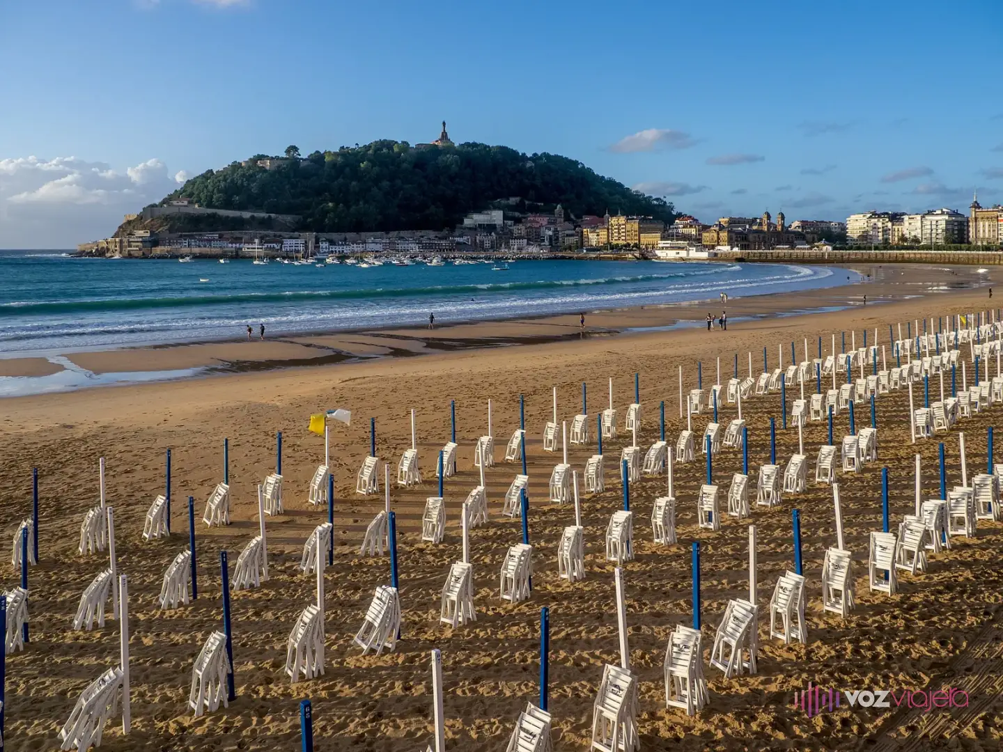 Filas de postes azules y blancos de los toldos, sin lona, y sillas blancas plegadas sobre la arena de La Concha al atardecer, con el monte Urgull al fondo