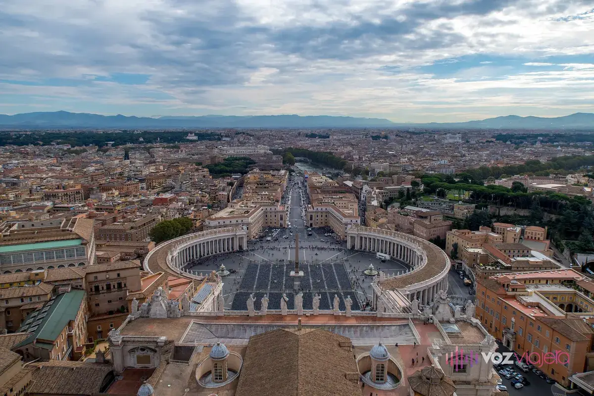 La plaza de San Pedro desde lo alto de la cúpula: las 2 columnatas curvas alrededor de la plaza ovalada, el obelisco en el centro y una avenida recta que lleva hacia el río y los tejados de Roma