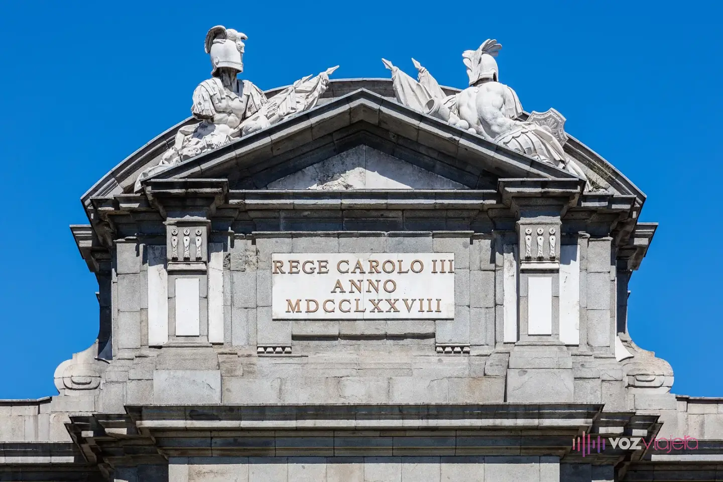Remate de la Puerta de Alcalá con dos grupos de trofeos militares de piedra con yelmos y banderas sobre un frontón y la lápida REGE CAROLO III ANNO MDCCLXXVIII