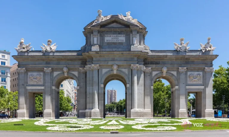 La Puerta de Alcalá de frente: arco de granito con 5 vanos, 3 arcos de medio punto en el centro y 2 vanos adintelados a los lados, trofeos militares en lo alto y un jardín delante