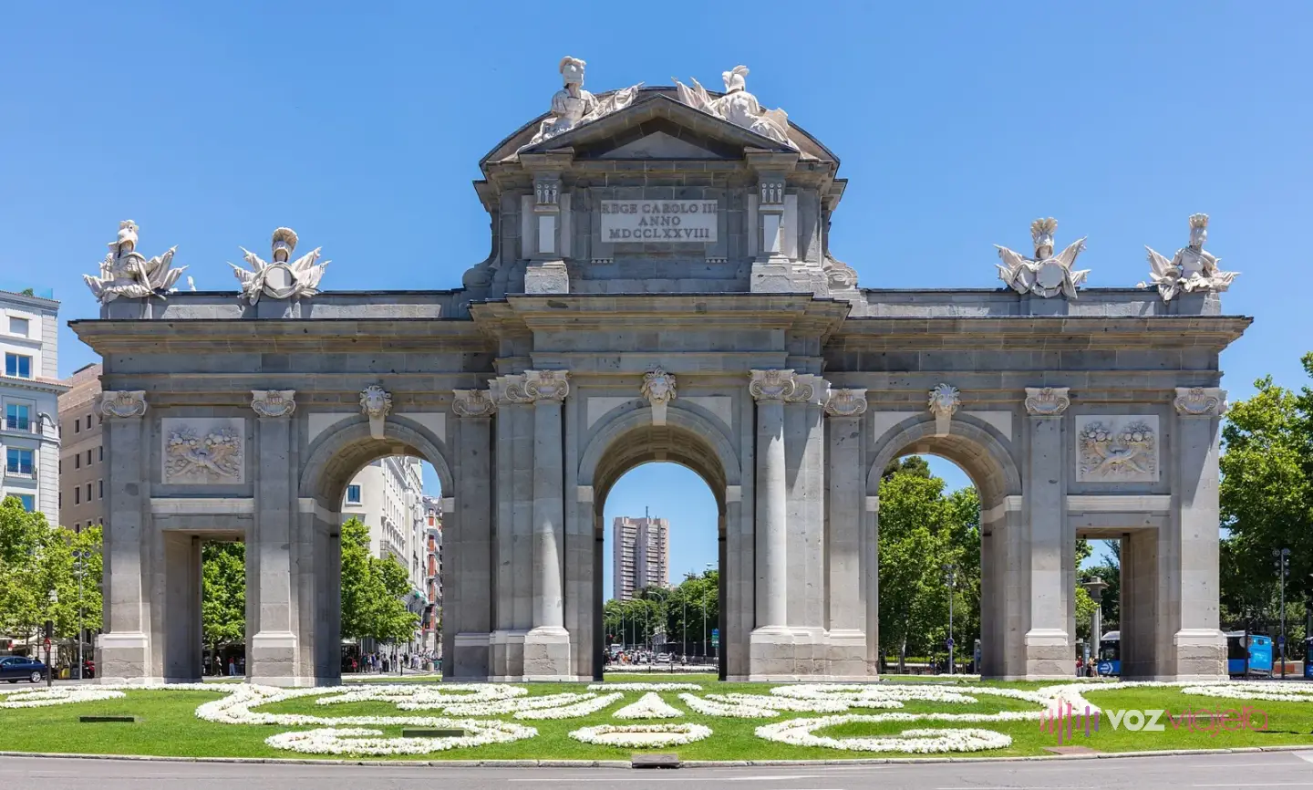 La Puerta de Alcalá de frente: arco de granito con 5 vanos, 3 arcos de medio punto en el centro y 2 vanos adintelados a los lados, trofeos militares en lo alto y un jardín delante