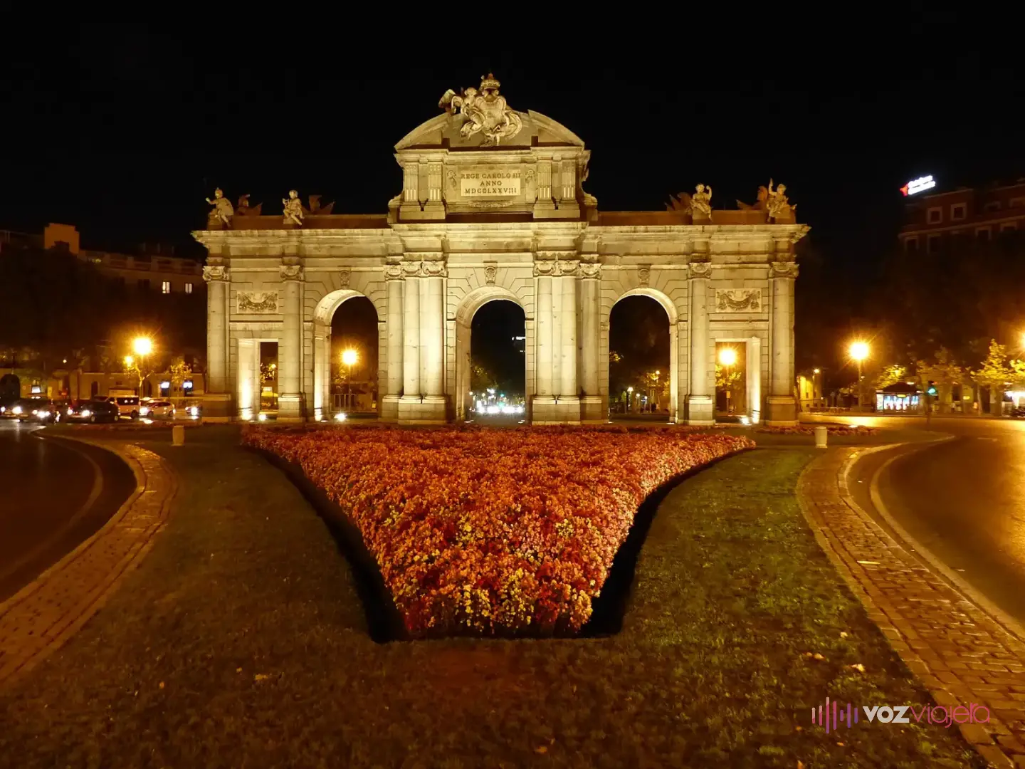 La Puerta de Alcalá iluminada de noche, con luz cálida sobre la piedra, farolas encendidas y un parterre de flores delante