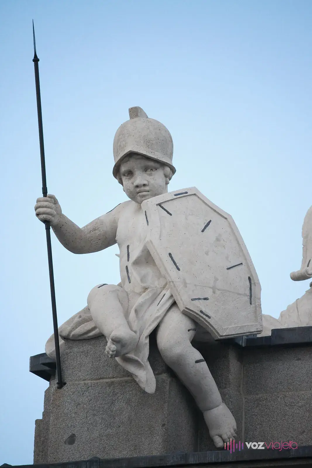 Escultura de piedra de un niño sentado con casco, escudo y una lanza de hierro: la Fortaleza, en lo alto de la Puerta de Alcalá