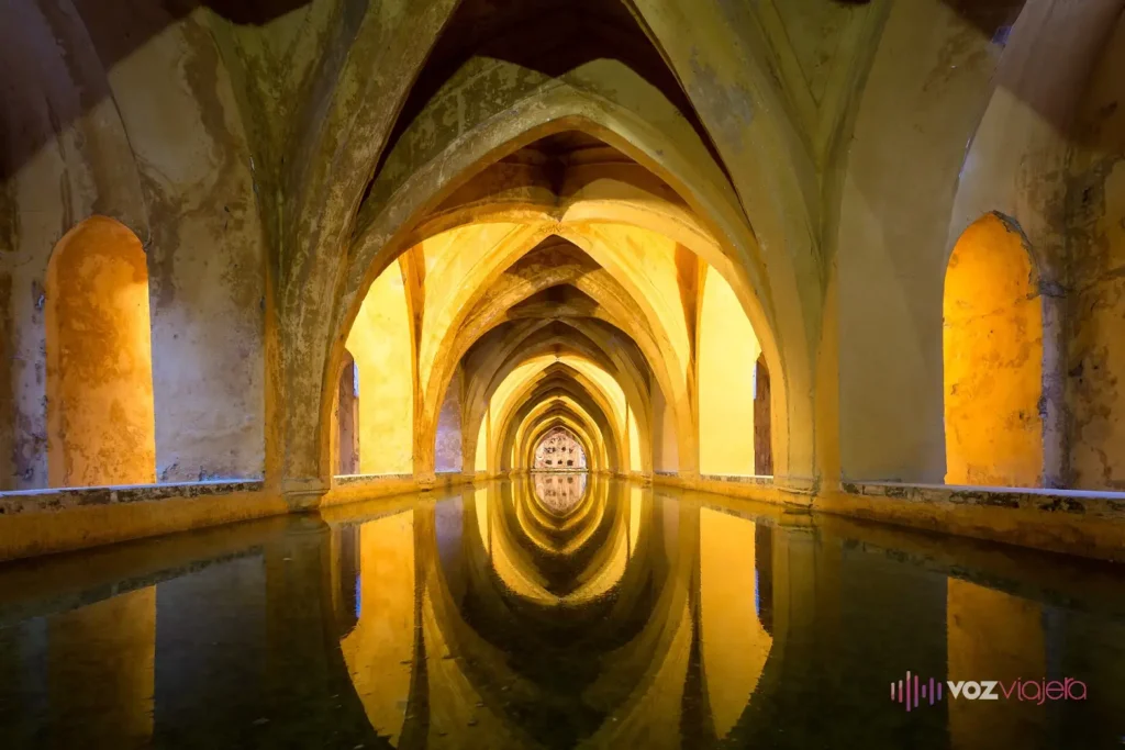 Los Baños de María de Padilla bajo el Real Alcázar de Sevilla: una galería alargada de bóvedas de arcos apuntados iluminada en tono cálido, reflejada en el agua quieta del estanque que ocupa el suelo