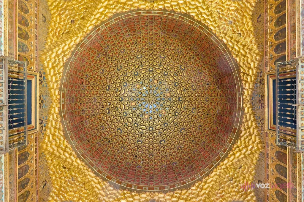 La cúpula dorada del Salón de Embajadores del Real Alcázar de Sevilla vista desde abajo: madera tallada y pintada en oro, rojo y azul, con un dibujo de estrellas, y pequeños balcones de madera a los lados