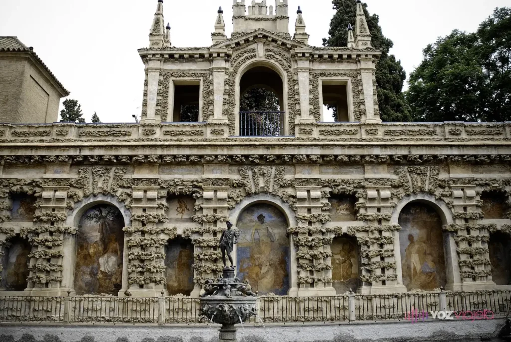 El Estanque de Mercurio del Real Alcázar de Sevilla con la estatua de bronce de Mercurio sobre su fuente, delante de la Galería del Grutesco, un muro de piedra rústica con hornacinas pintadas y un mirador en lo alto