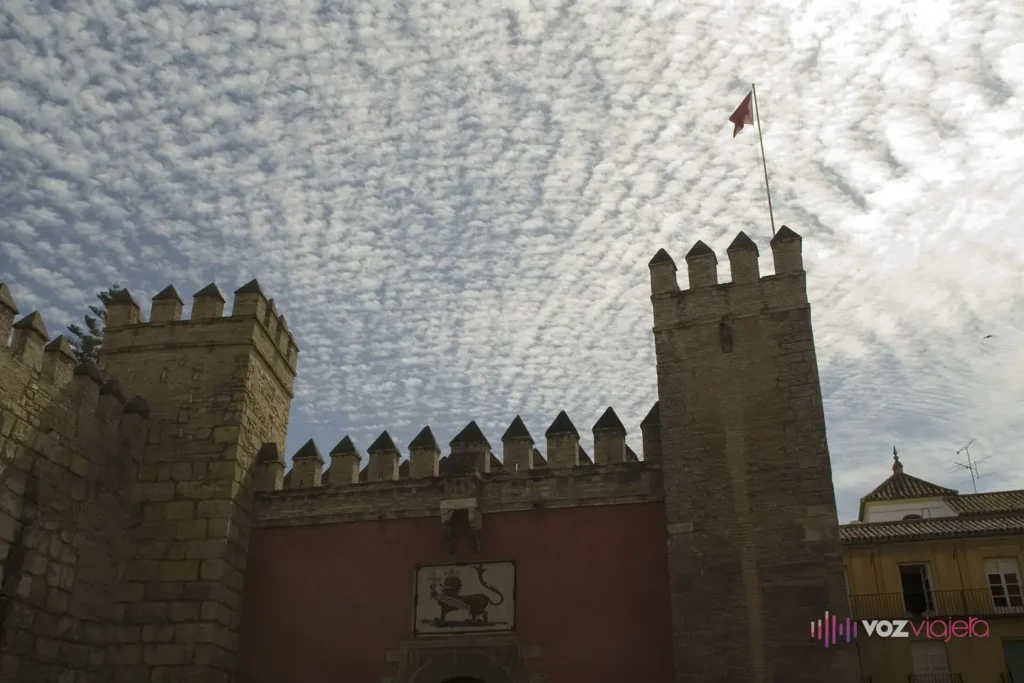 La Puerta del León del Real Alcázar de Sevilla: un paño pintado de rojo entre 2 torres almenadas, con un panel de azulejos de un león coronado sobre la puerta y una bandera en lo alto de la torre