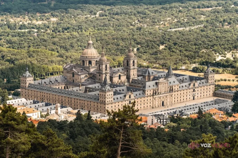 Vista general del Real Monasterio de El Escorial, un gran edificio de granito con cúpula y torres, rodeado de arbolado y tejados de la villa